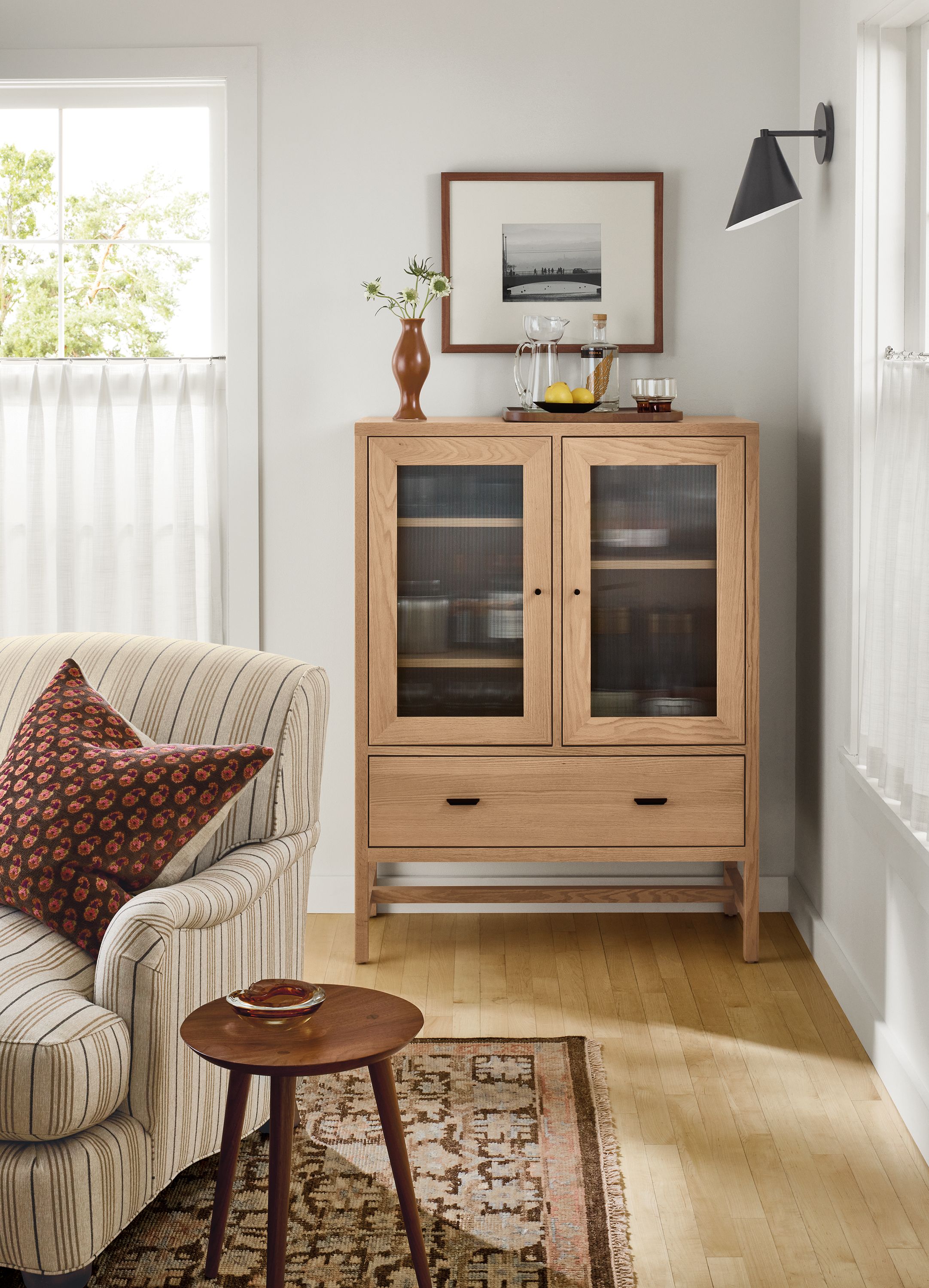 Living room corner with Berkeley linen cabinet with glass door in rye on red oak, Humphrey chair in Prescott natural and Rosalie rug in petal.
