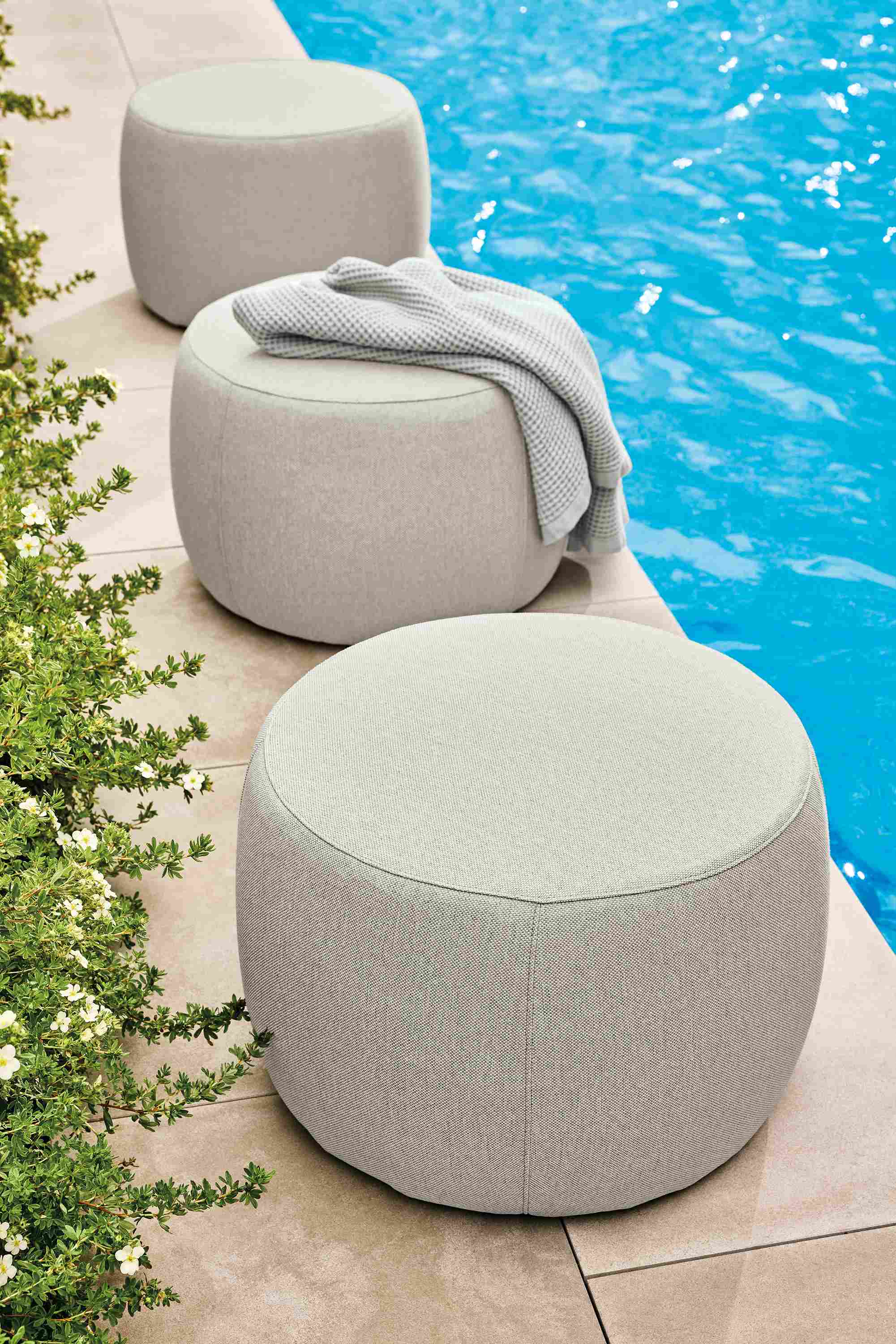 Poolside view of 3 Boyd round ottomans in Mist Grey and a Lawrence bath towel in grey.