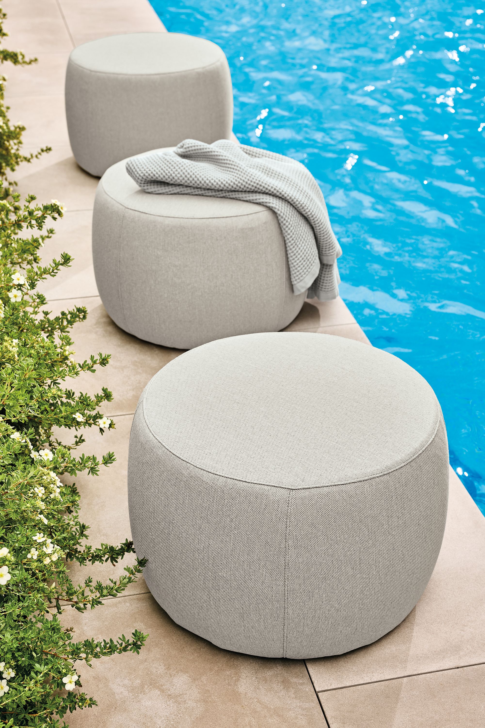 Poolside view of 3 Boyd round ottomans in Mist Grey and a Lawrence bath towel in grey.