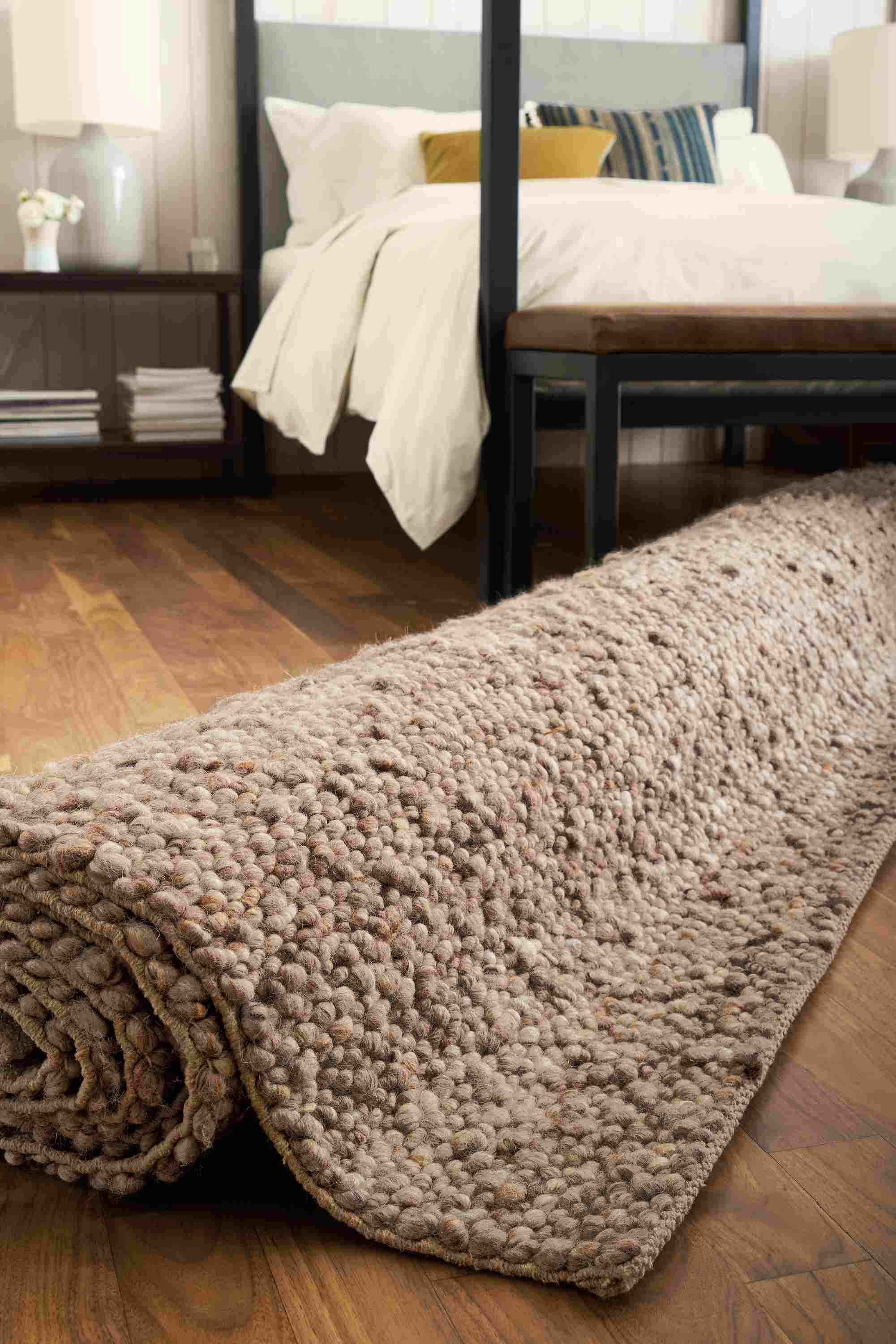 Low-angle view of a rolled-up cobble rug in putty, architecture queen bed in derry and serena table lamp in grey.