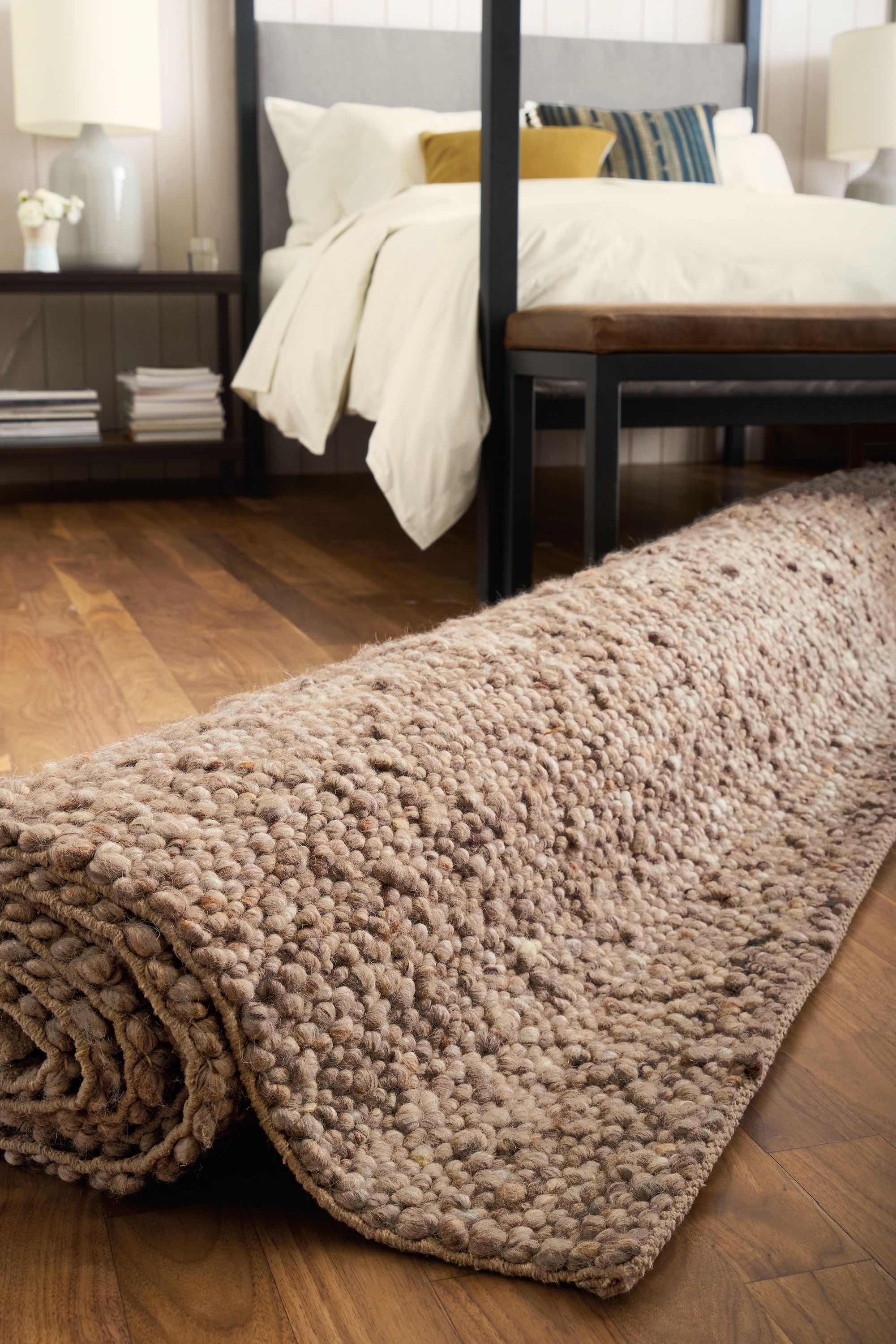 Low-angle view of a rolled-up cobble rug in putty, architecture queen bed in derry and serena table lamp in grey.