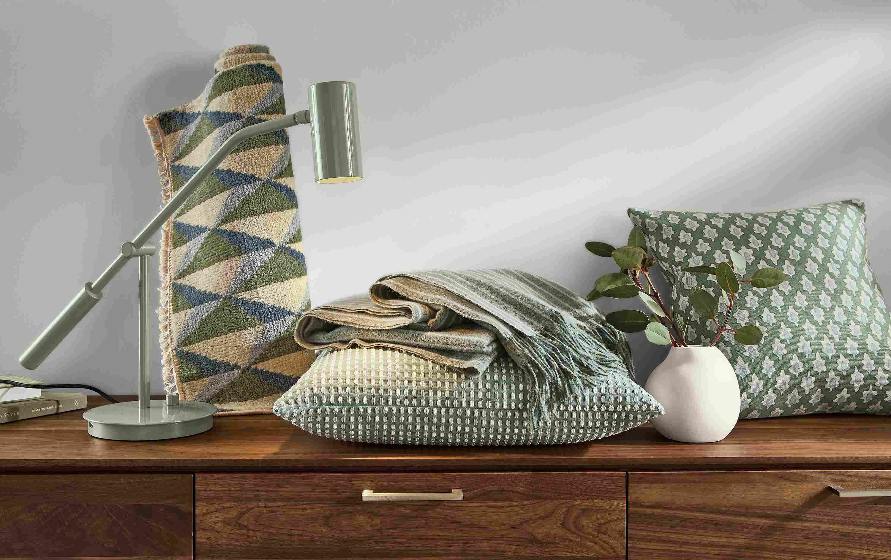 Green items like rug, lamp, pillow and throw blanket sitting on top of walnut dresser.