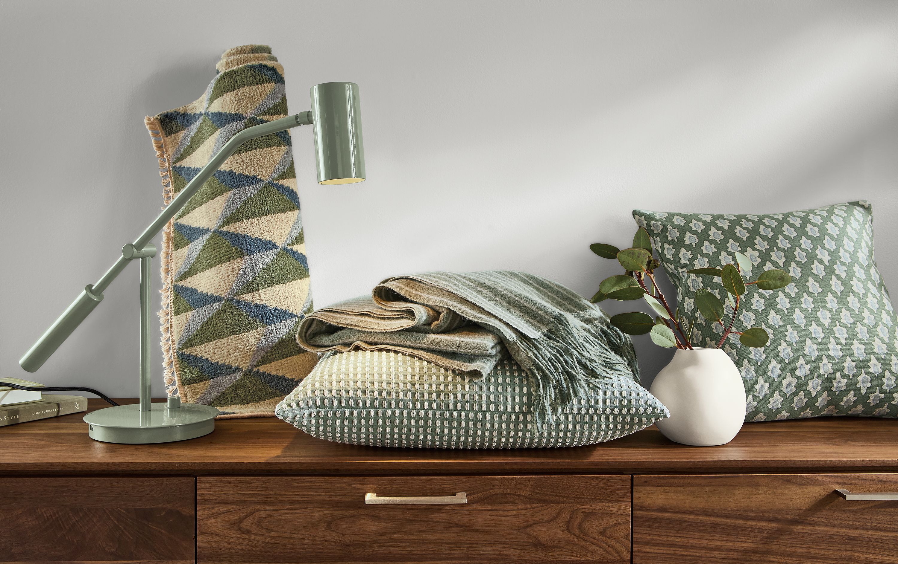 Green items like rug, lamp, pillow and throw blanket sitting on top of walnut dresser.