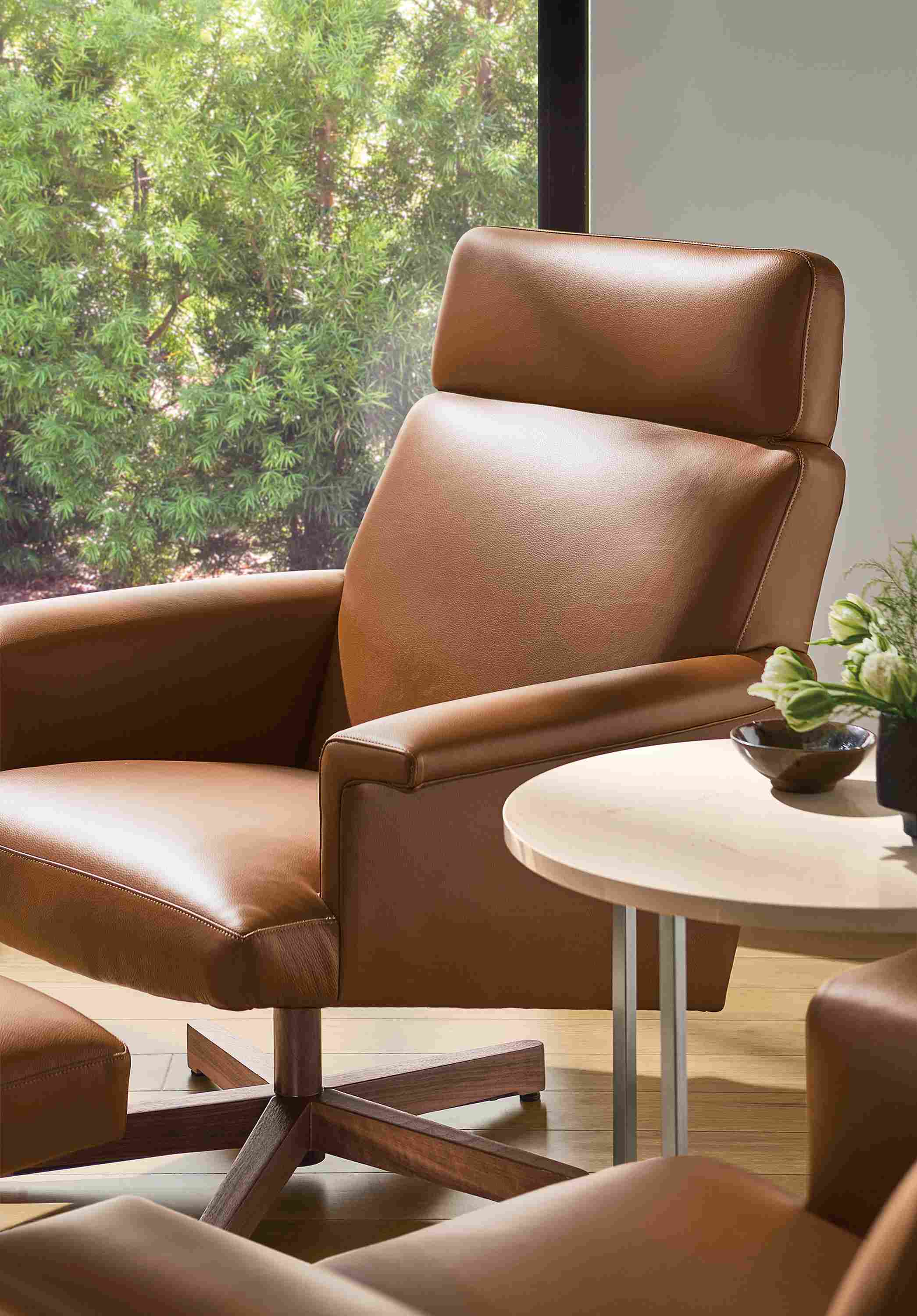 Detail of daniels swivel chair and ottoman in pesaro cognac leather and walnut and classic round table in ecru quartz.
