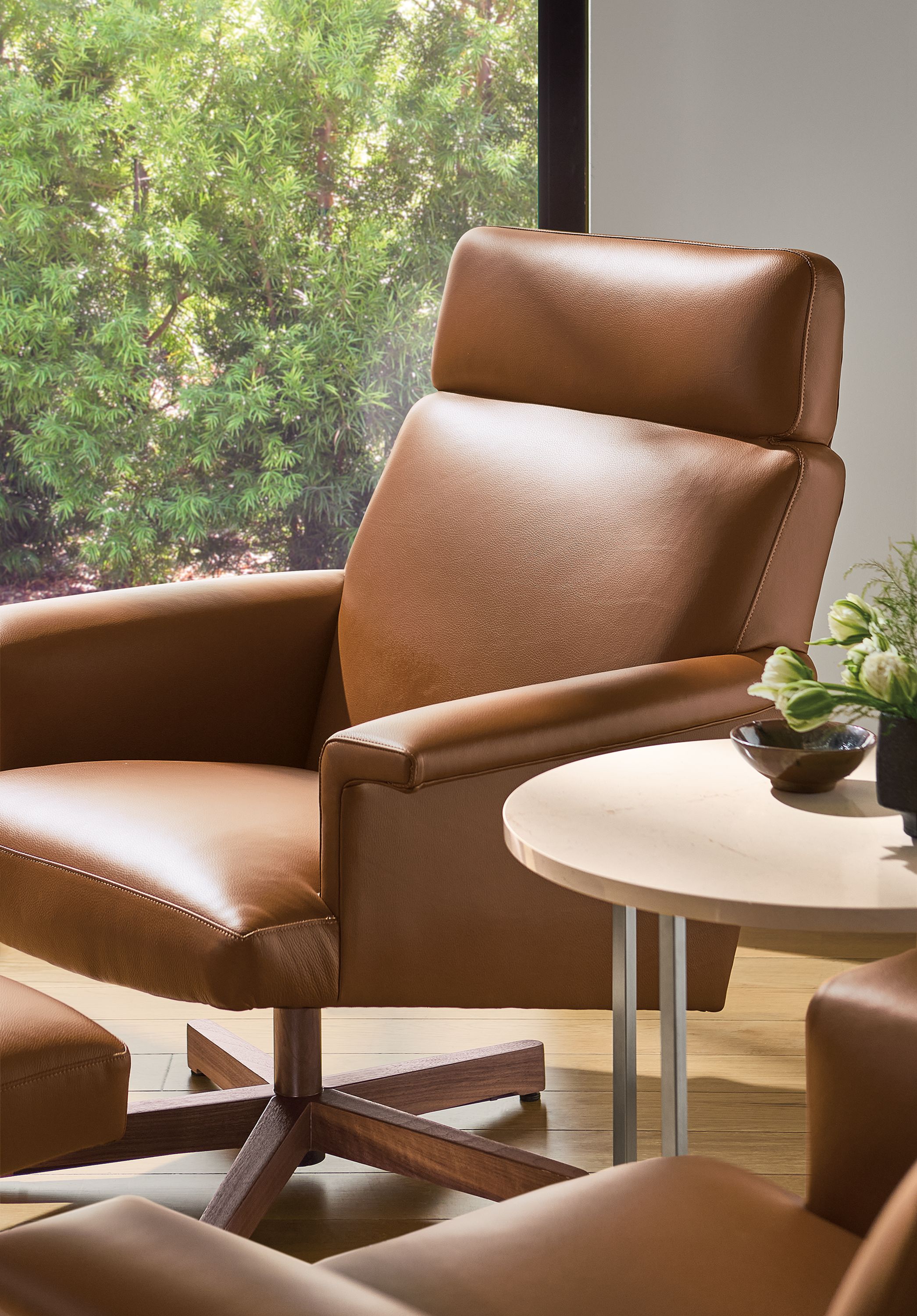 Detail of daniels swivel chair and ottoman in pesaro cognac leather and walnut and classic round table in ecru quartz.