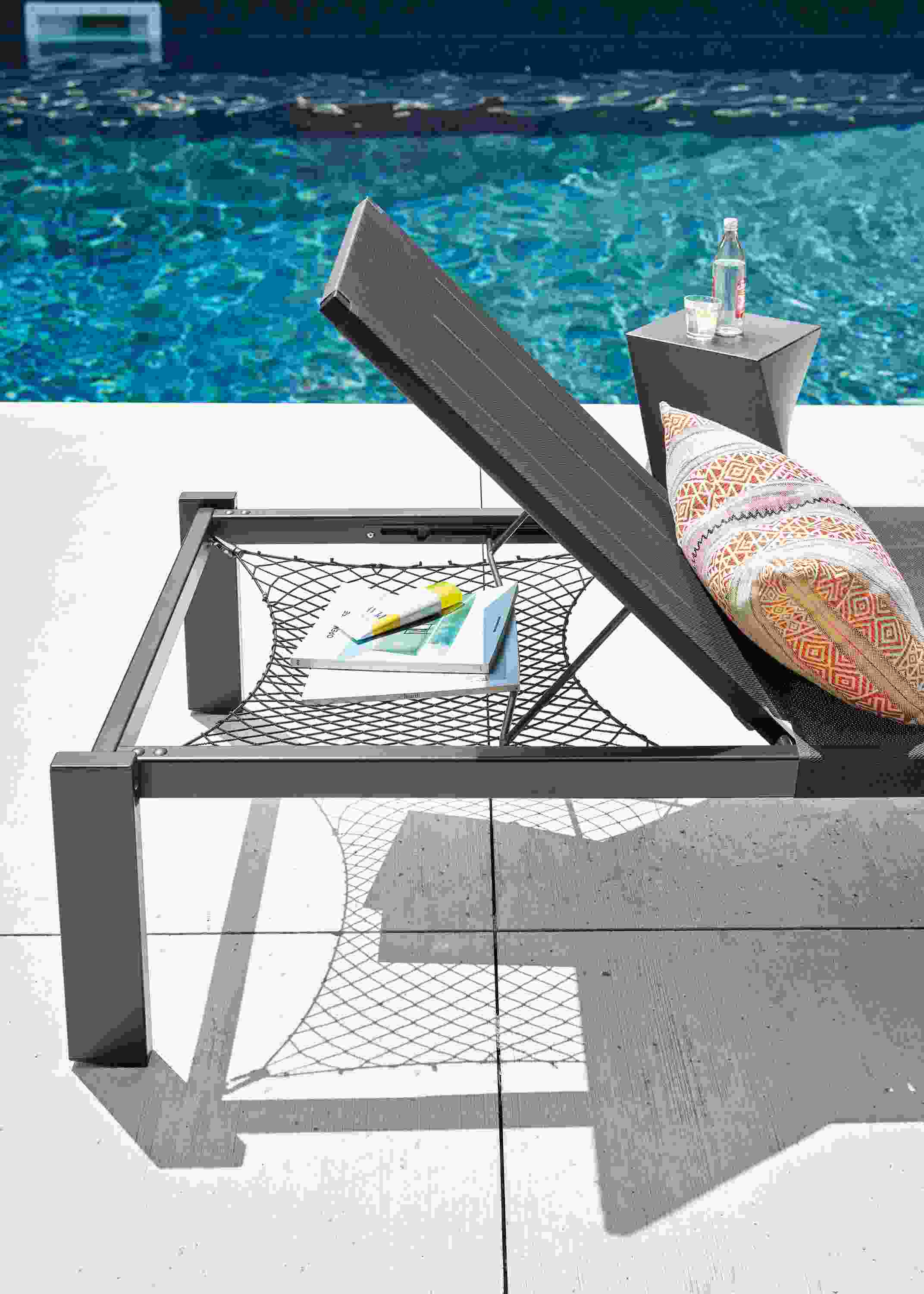Poolside detail of a Dorian chaise with storage net in black showing the storage net in use holding books and sunscreen.