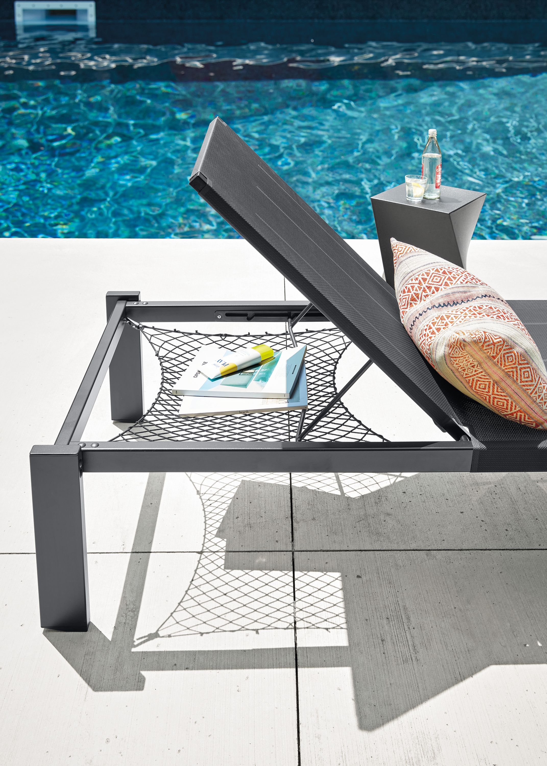 Poolside detail of a Dorian chaise with storage net in black showing the storage net in use holding books and sunscreen.