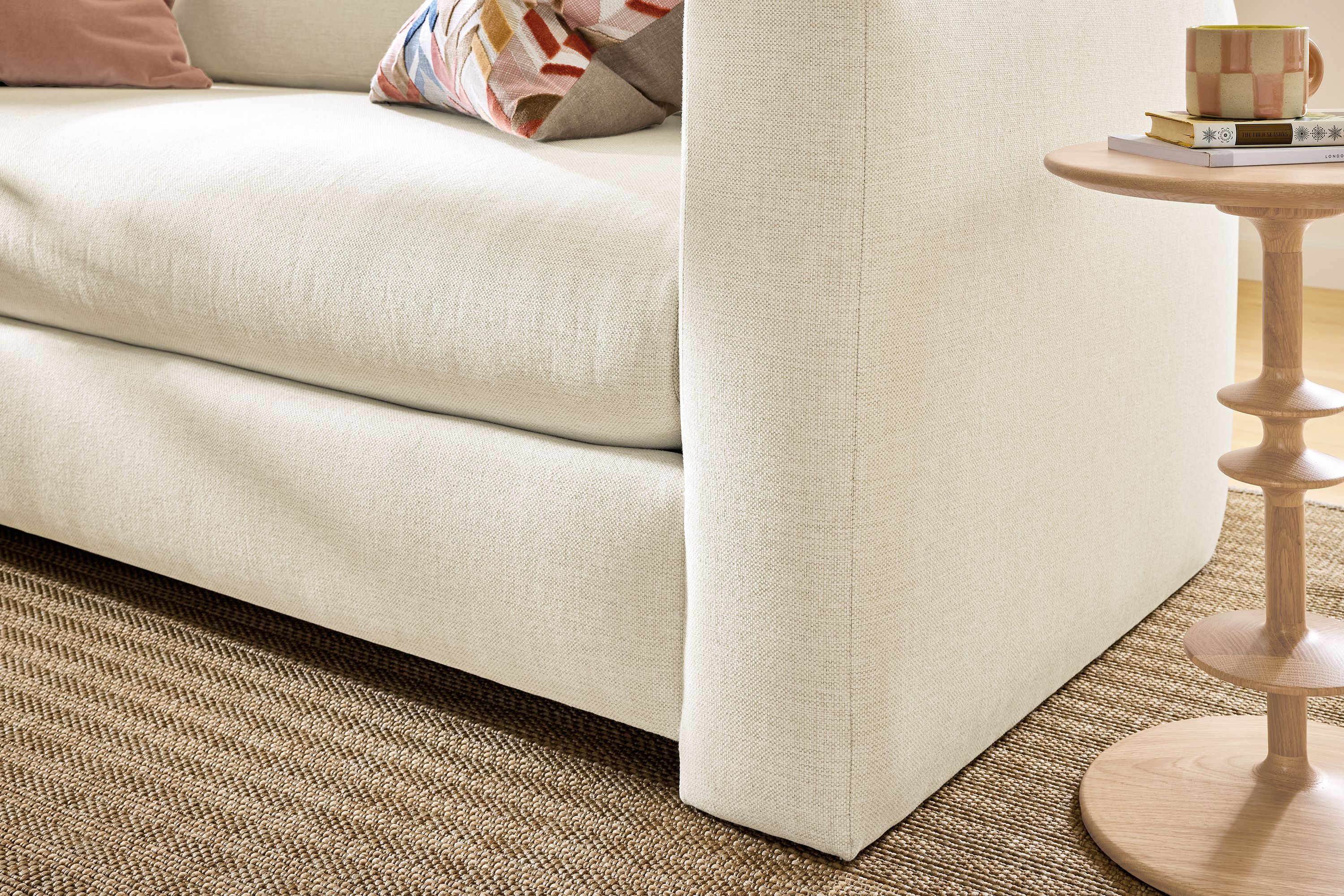 Detail of the front corner of a Hannah 76-wide bench cushion sofa in sumner ivory and Leland rug in sand in a living room.