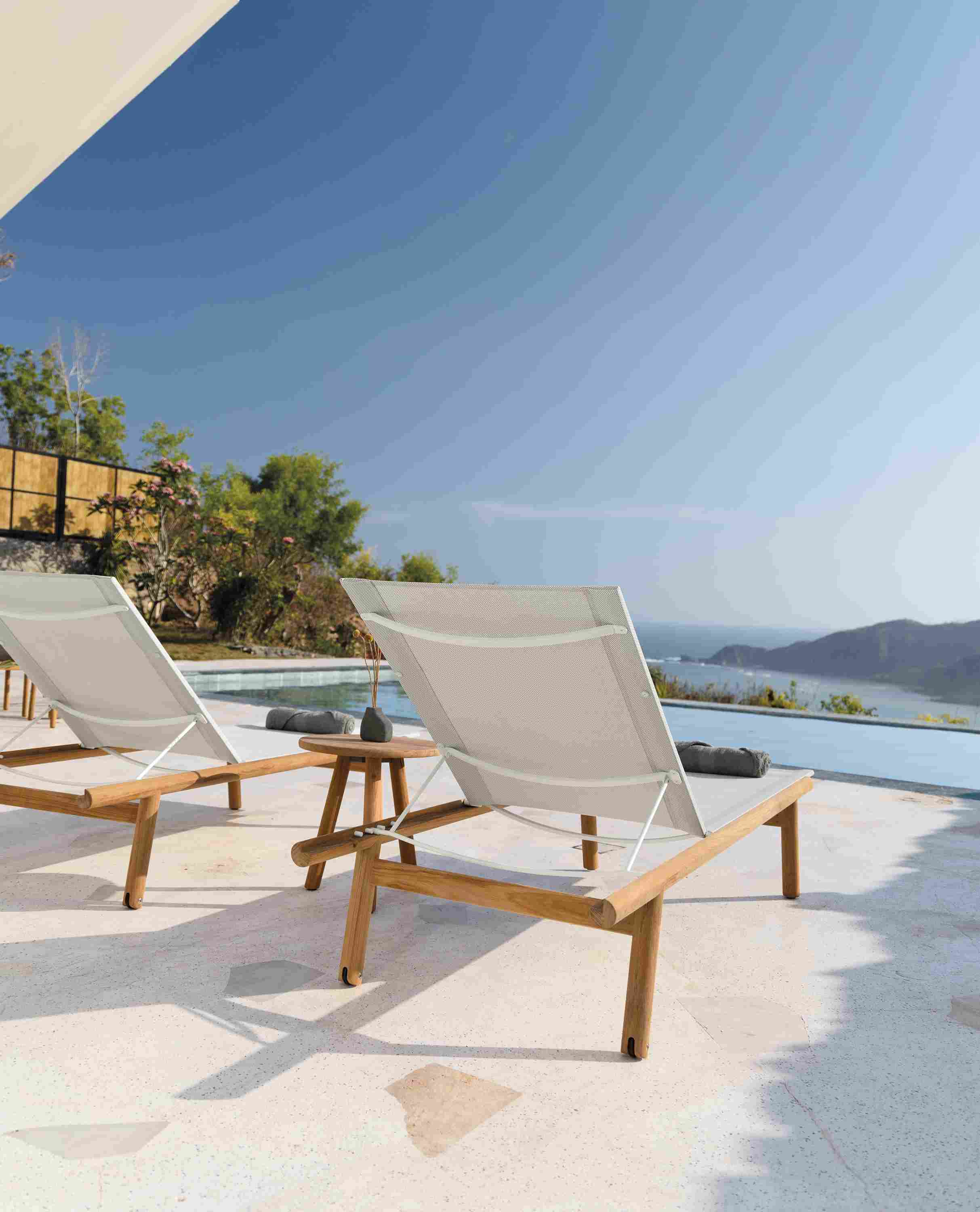 Back view of two Isley Chaises in Teak with Salt Sling with a 17-round Isley table.