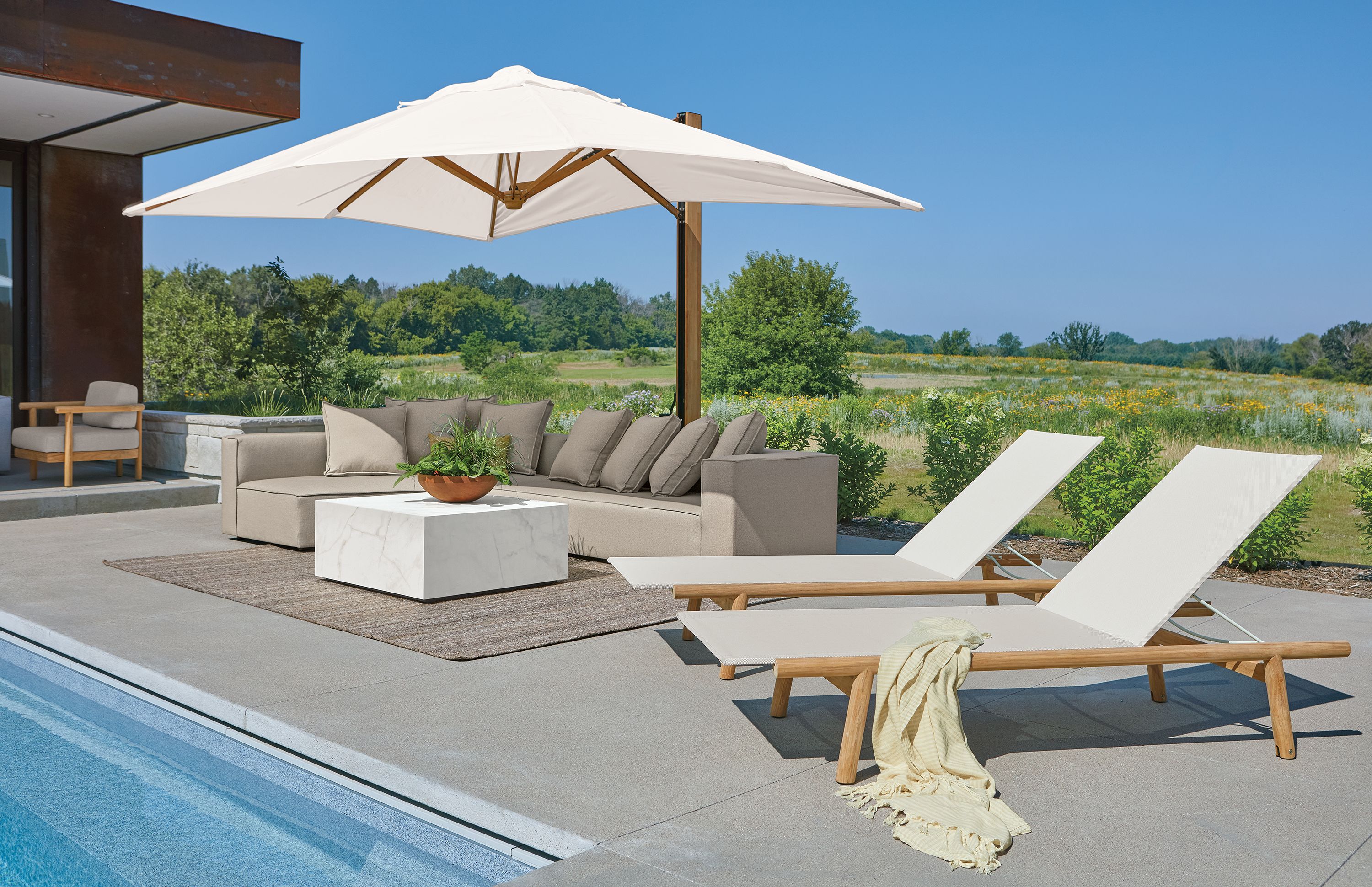 Poolside area with 2 Isley chaises in teak with salt sling and Banda cantilever patio umbrella in teak and halo white.