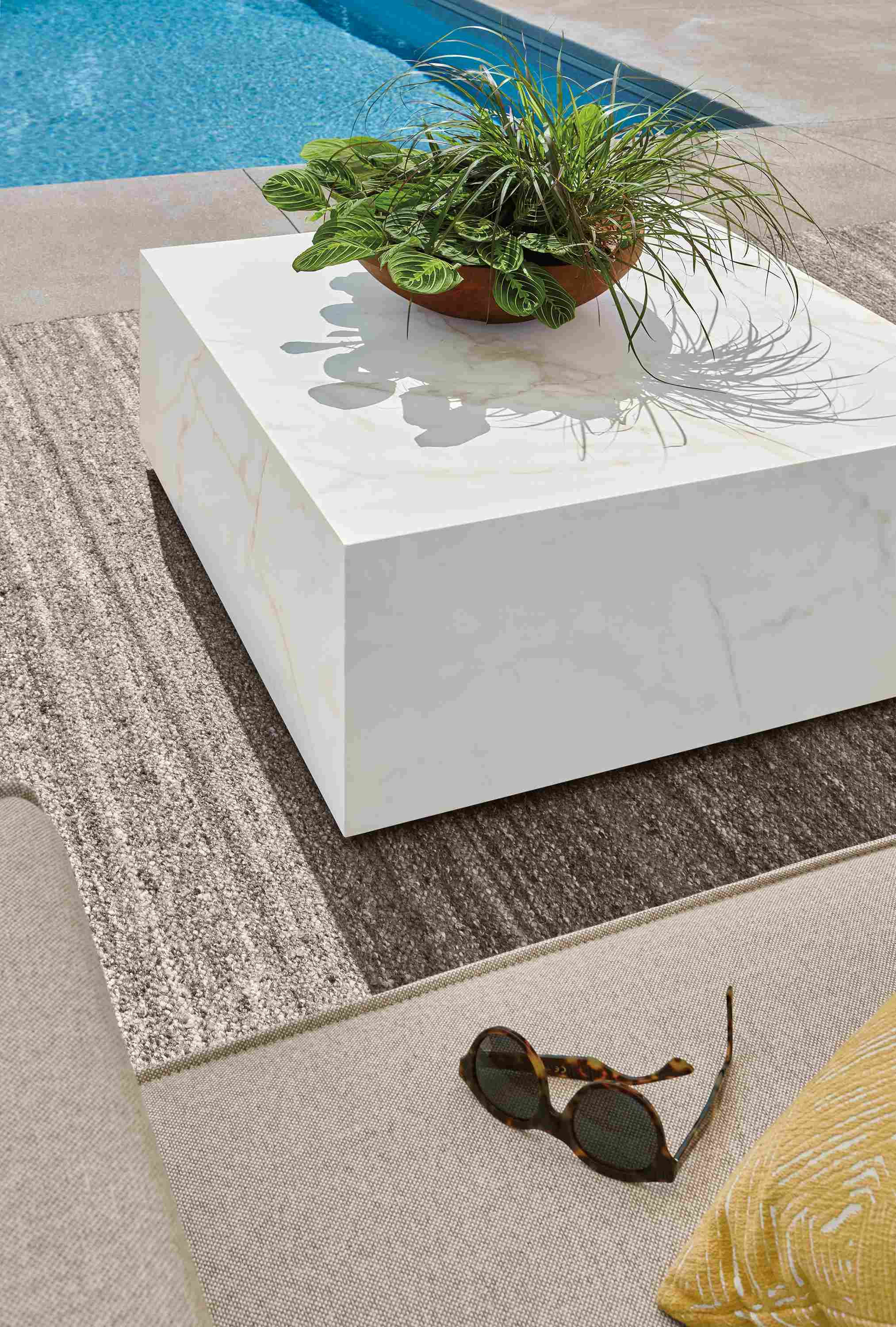 Poolside view of Leona 36 by 36 outdoor coffee table in marbled white/taupe ceramic and Sefton rug in earth.