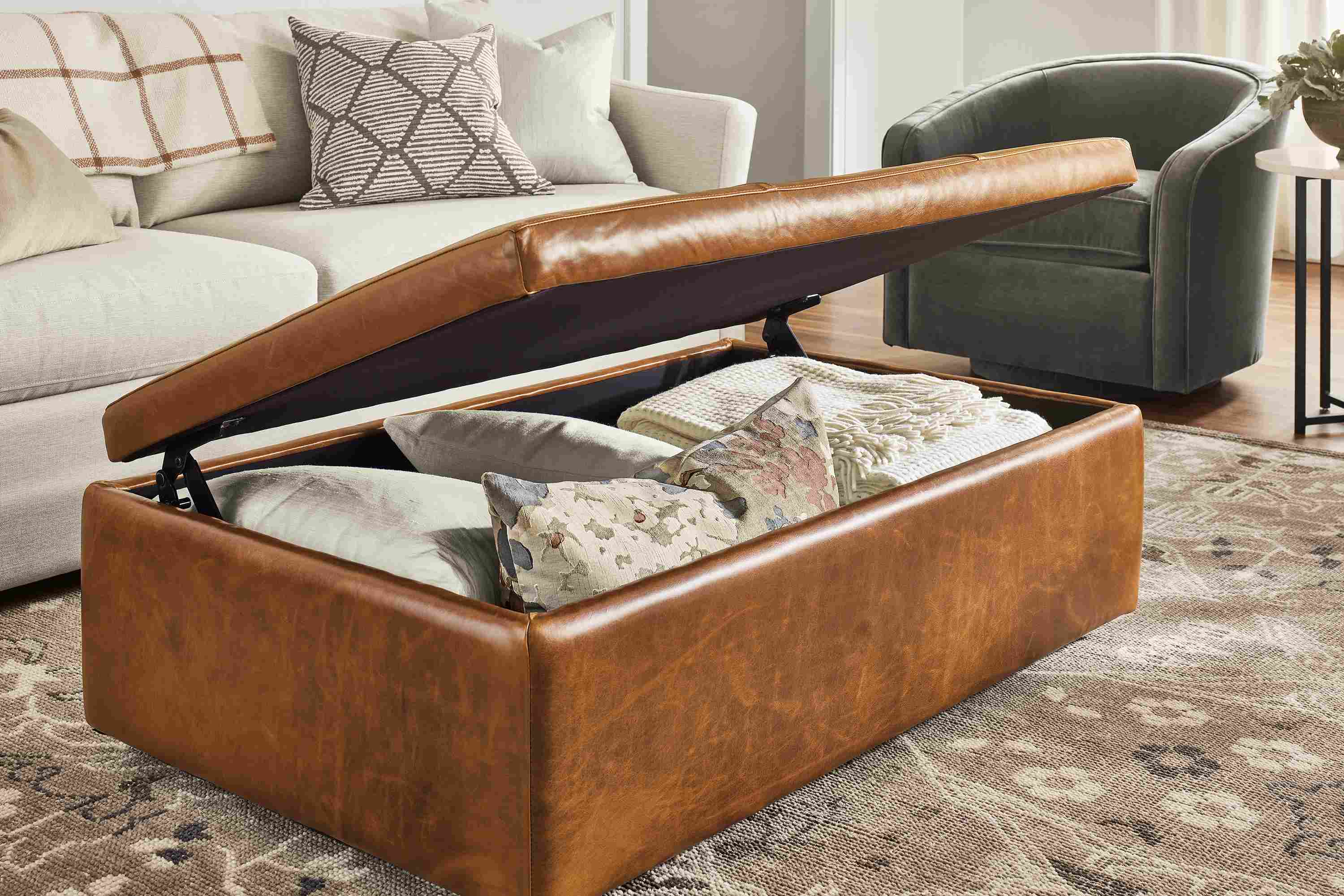 Leather Lind storage ottoman in Vento Cognac with lid open showing throw pillows and blankets inside.