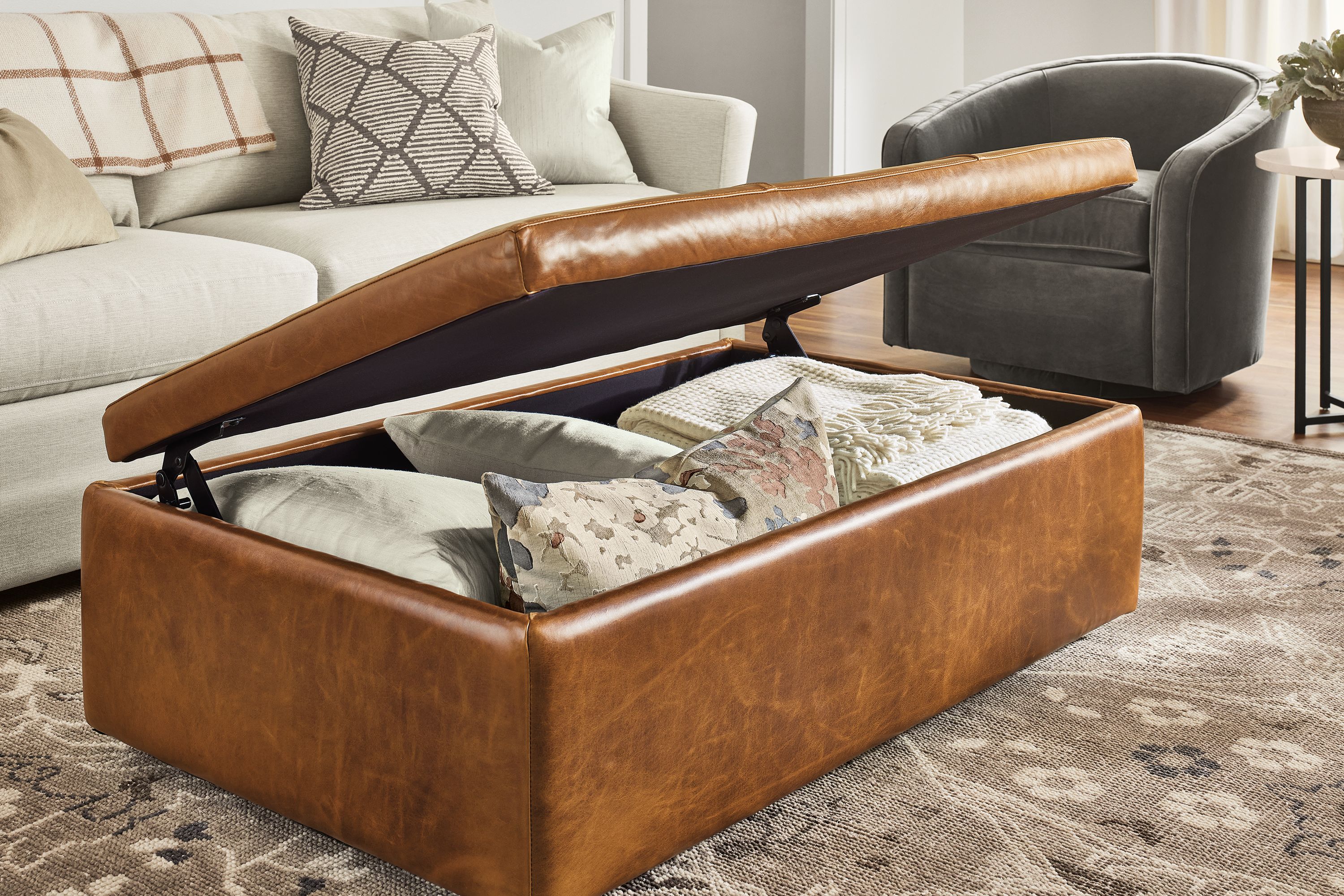 Leather Lind storage ottoman in Vento Cognac with lid open showing throw pillows and blankets inside.
