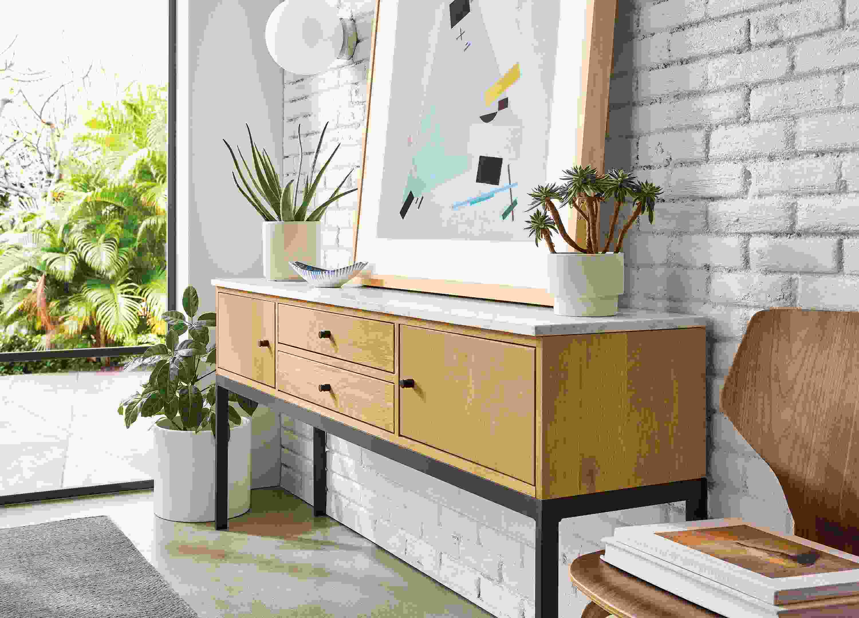linear 72-wide storage cabinet in white oak with quartz top, with leaning wall art and houseplants.