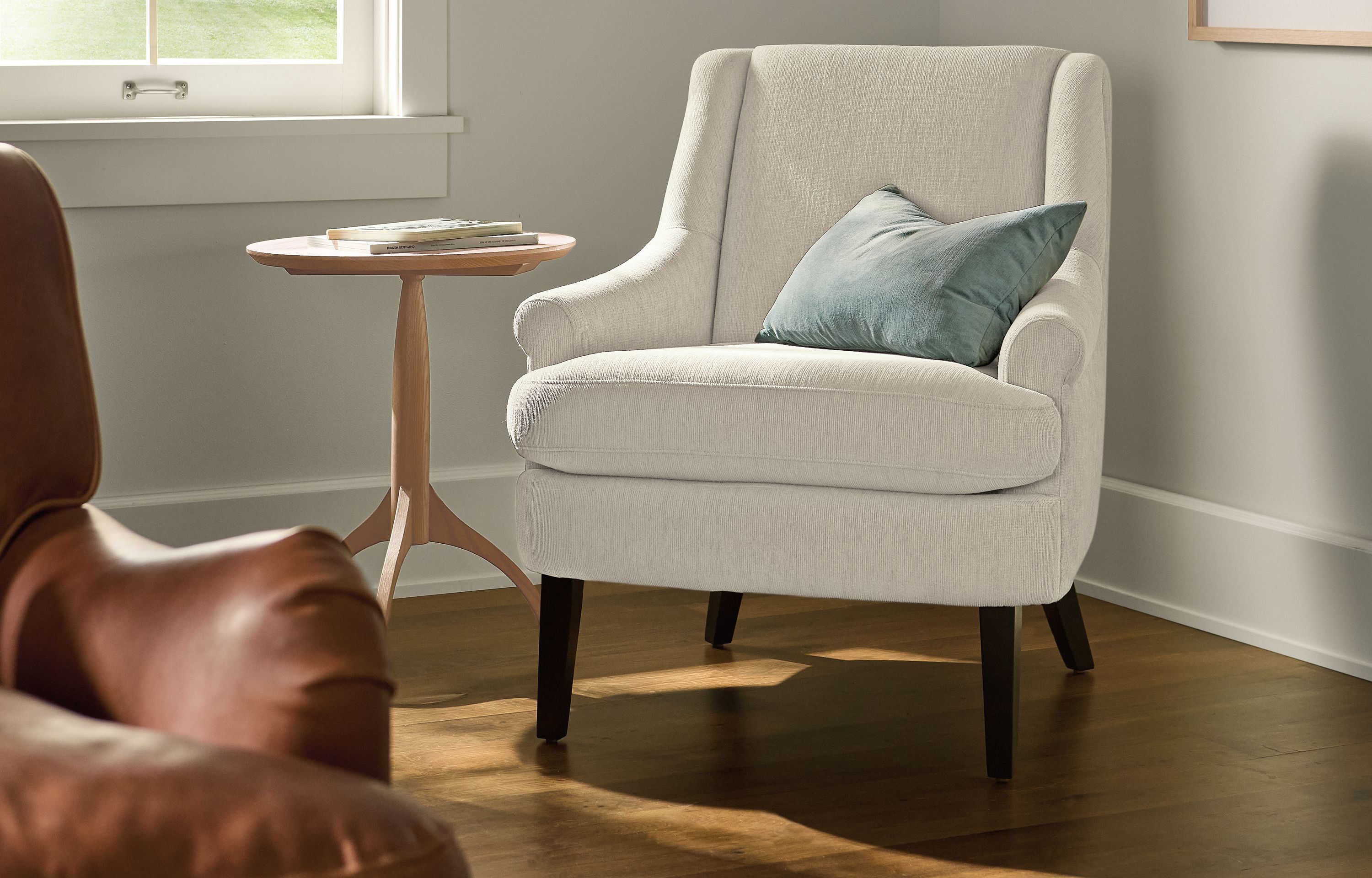 Living room corner with a Louise chair in Crawford Bone, Adams round table in rye on red oak and Le Beuan Benic wall art.
