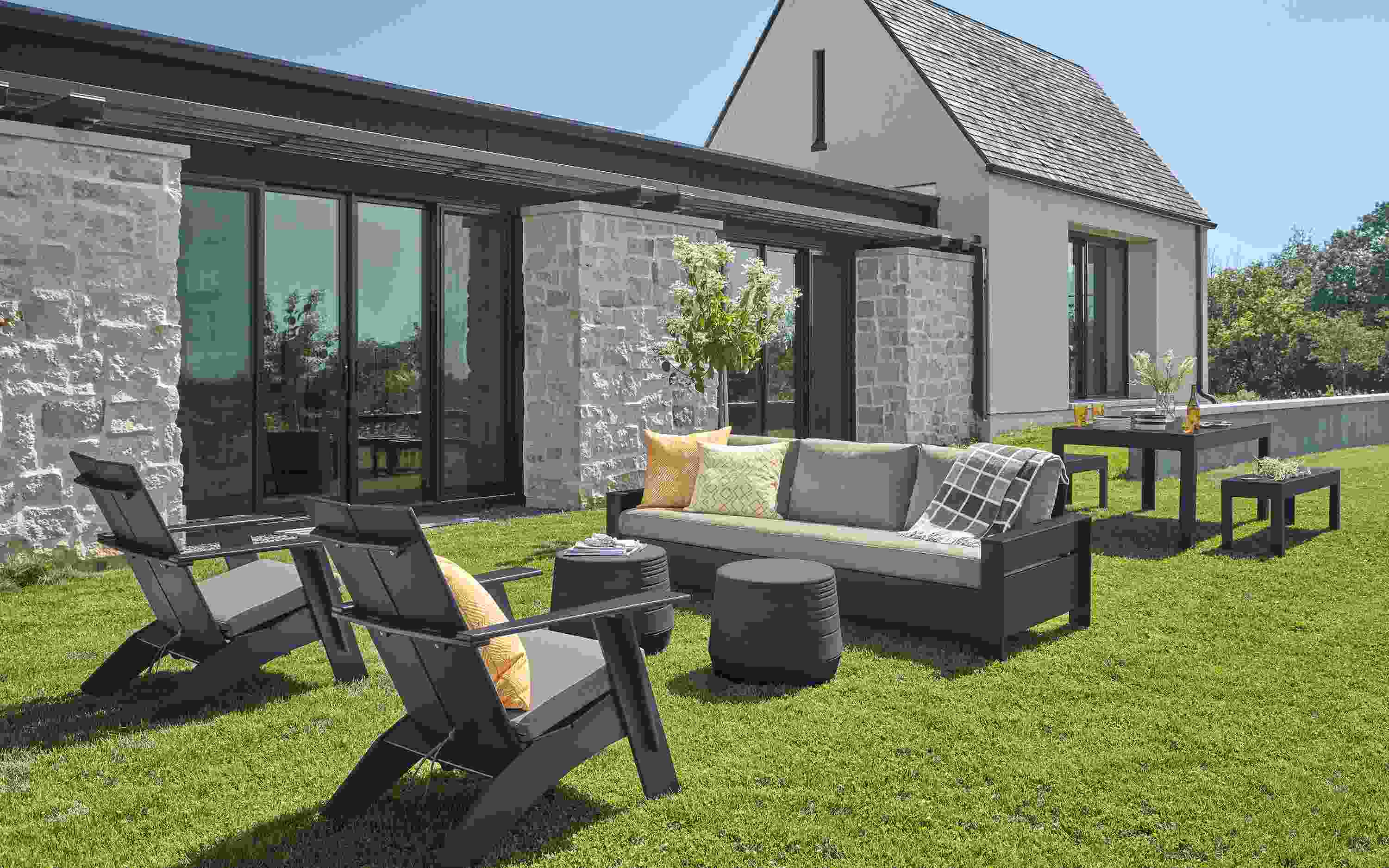 Rayo Sofa and Emmet Tall Lounge Chairs in black with Pelham charcoal cushions and Cusp Stool in Grey on a lawn.