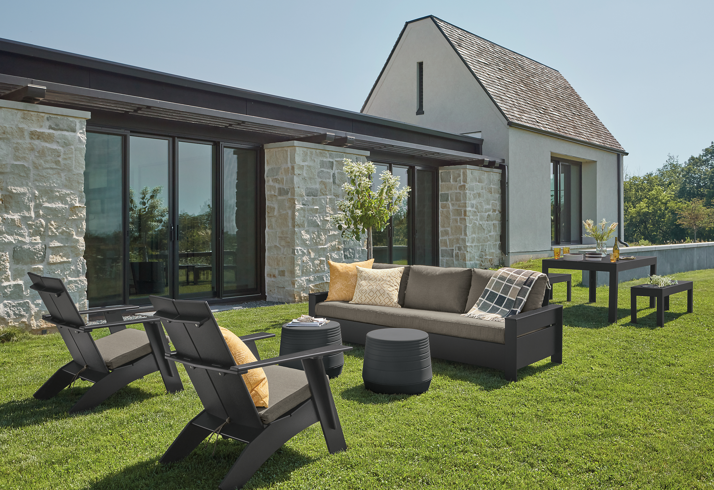Rayo Sofa and Emmet Tall Lounge Chairs in black with Pelham charcoal cushions and Cusp Stool in Grey on a lawn.