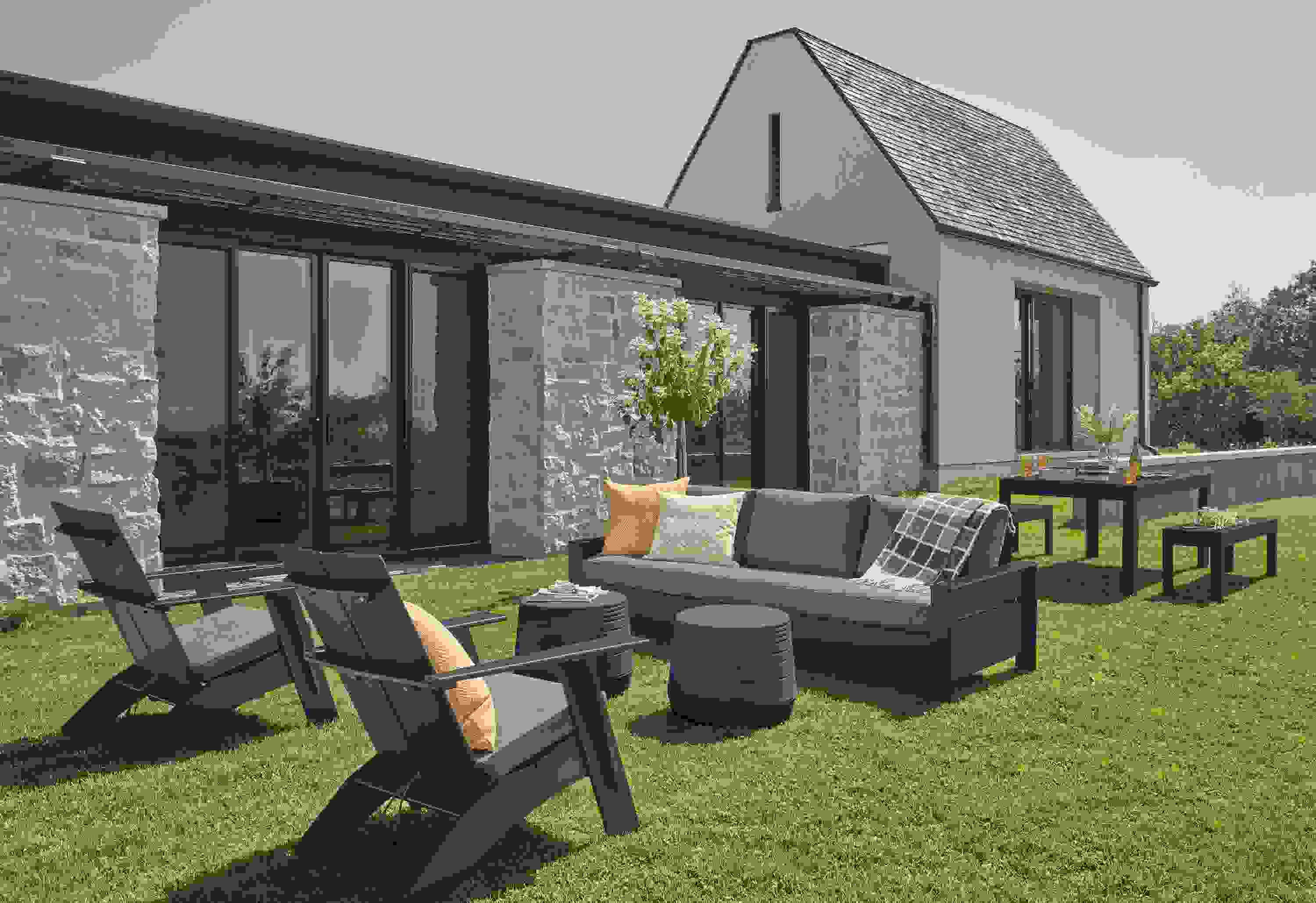 Rayo Sofa and Emmet Tall Lounge Chairs in black with Pelham charcoal cushions and Cusp Stool in Grey on a lawn.